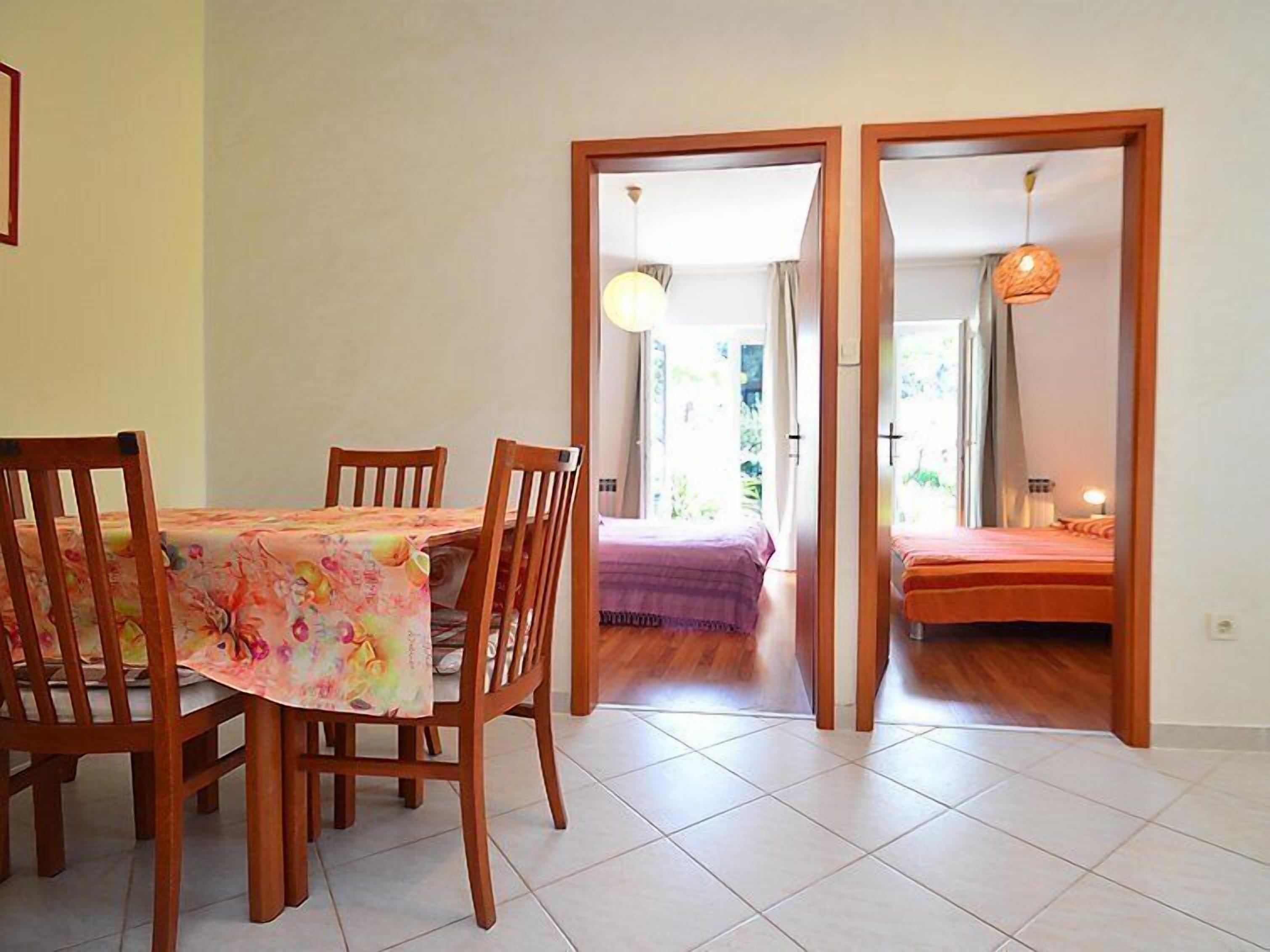 Zona pranzo con tavolo e sedie, un letto con coperta viola e vista su un giardino attraverso una porta aperta.