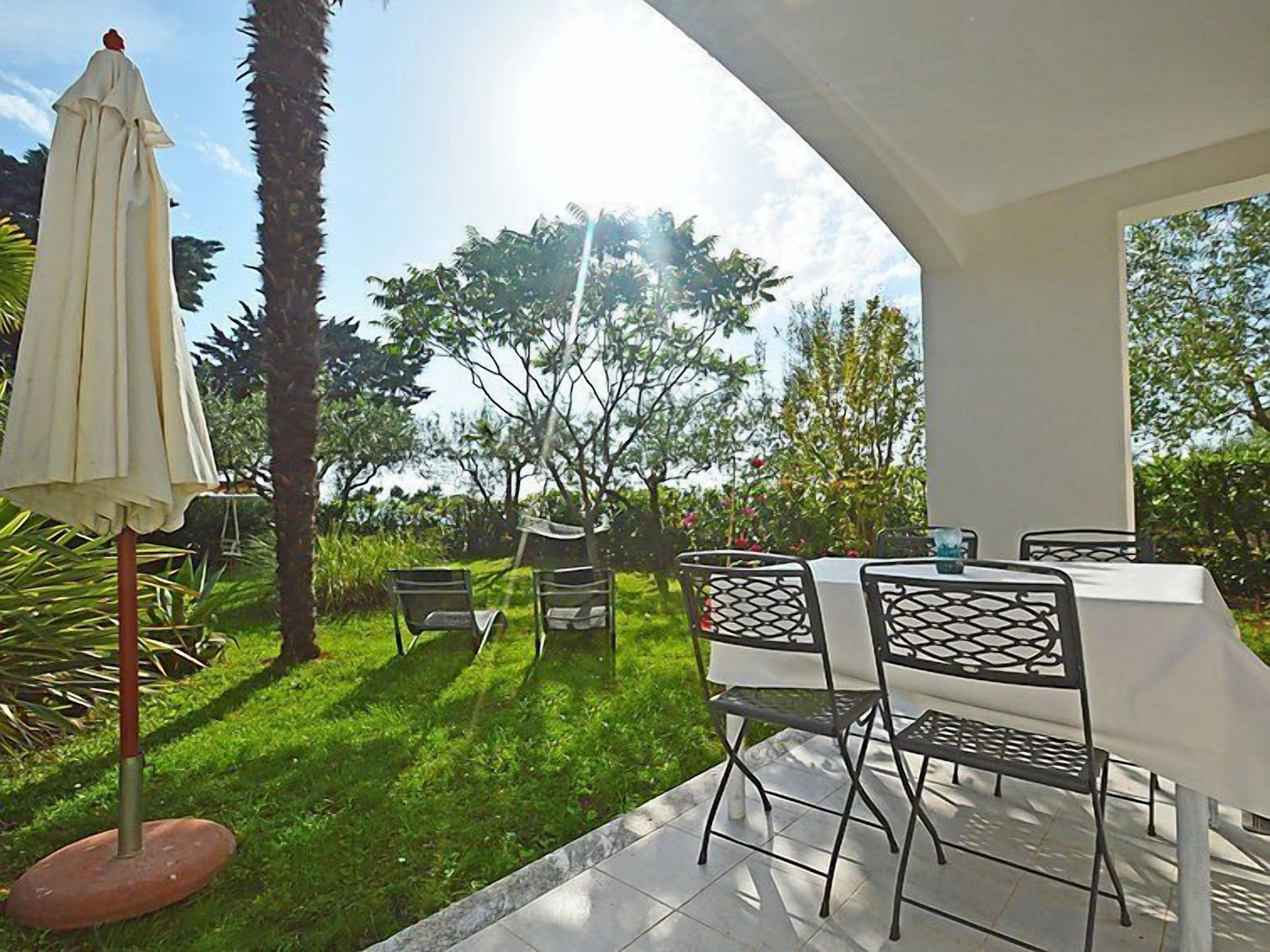 Eine sonnige Terrassenanlage mit Tisch und Stühlen, umgeben von Pflanzen und Bäumen.