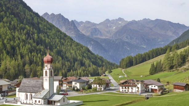 A rural village with a church featuring a prominent red dome, surrounded by houses and fields, with mountains in the background.