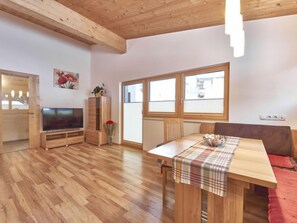A spacious living room with wooden flooring, a TV, a wooden cabinet, a dining table with a plaid tablecloth, and large windows.