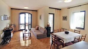 Un salon spacieux comprenant un canapé, une table à manger avec des chaises, un parquet en bois à motifs chevrons et une grande fenêtre avec des rideaux.
