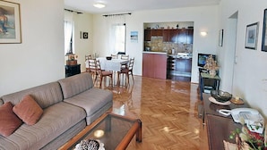 Un salon comprenant un canapé, une table basse et un coin repas avec des chaises et une table. On aperçoit une cuisine au fond.