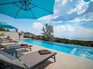 Uma área à beira da piscina com espreguiçadeiras, um guarda-sol azul e vista para o mar.