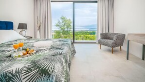 Ein modernes Hotelzimmer mit einem großen Bett, einem Sessel, einem Schreibtisch und einem Balkon mit Blick auf Grünflächen und das Meer.