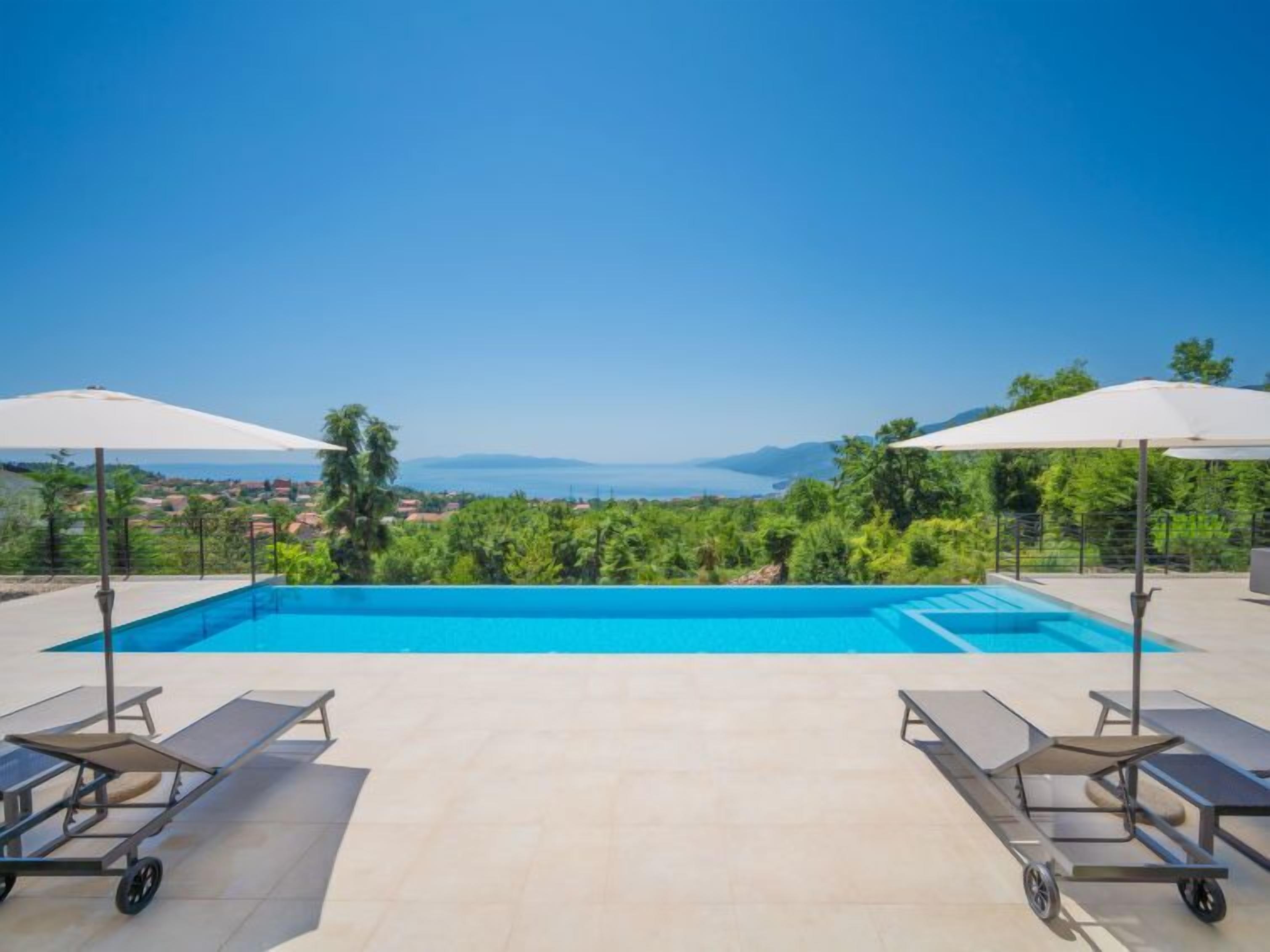 A rectangular swimming pool with a clear blue water, surrounded by a tiled deck. There are two lounge chairs with sun umbrellas on either side of the pool.