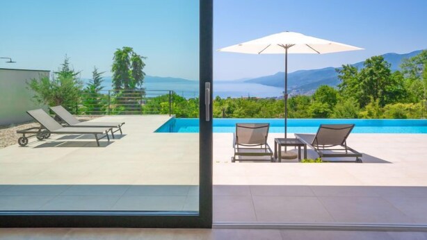 Ein moderner Poolbereich im Freien mit Liegestühlen und einem Sonnenschirm, mit Blick auf eine malerische Aussicht auf Meer und Berge.
