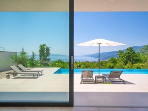 Un área de piscina exterior moderna con sillones de descanso y una sombrilla, con vistas panorámicas al mar y las montañas.