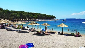 A beach with umbrellas and lounge chairs, a clear blue sea, and a forested hill in the background.