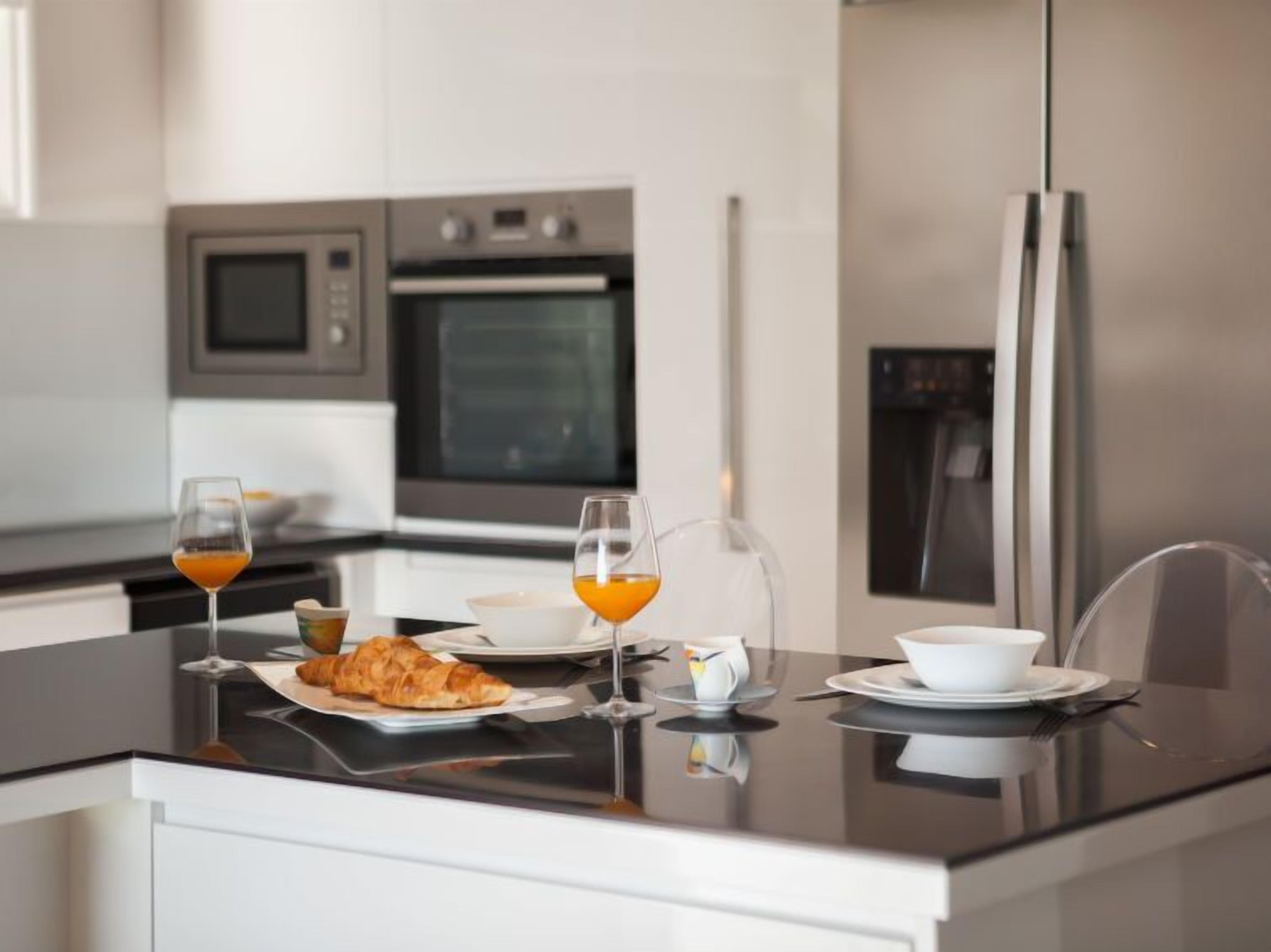Une cuisine moderne équipée d’appareils électroménagers en acier inoxydable, une table à manger en verre et un assortiment de produits pour le petit-déjeuner, notamment des croissants, du jus de fruits et une théière.