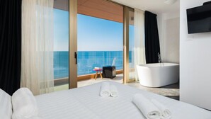 A hotel room with a bed, a small table with a chair, a bathtub, and a large window with a view of the sea.