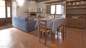 Un comedor rústico con una mesa de madera dispuesta para cuatro personas, rodeada de sillas de madera. Un sofá está situado contra la pared, y se observa una chimenea al fondo.