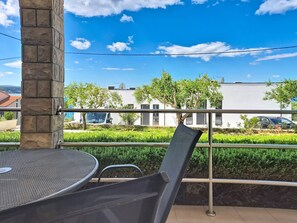 En balkon med en metalstol, et bord og udsigt til en hvid bygning med parkerede biler og træer.