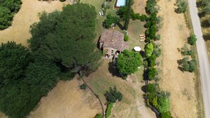Vista aérea de uma propriedade rural com um edifício central rodeado por árvores densas, uma piscina pequena e uma estrada pavimentada.