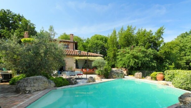 A swimming pool with a tiled deck, surrounded by rocks and greenery, and a house with a tiled roof in the background. - Vacation home Oasis by Interhome (Penna in Teverina)