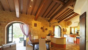 A rustic stone interior with a wooden ceiling, a dining area with white chairs, a kitchen with a counter, and a large window with curtains. - Vacation home Oasis by Interhome (Penna in Teverina)