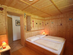 A wooden cabin bedroom with two beds, a nightstand with a lamp, and a door leading to another room.