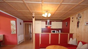 Une pièce chaleureuse avec un plancher en bois, une table ronde en bois, un canapé rouge avec des coussins blancs, une kitchenette avec des meubles blancs et un plafond avec des poutres en bois et un luminaire suspendu.