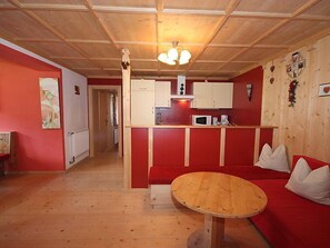A cozy room with a wooden floor, a round wooden table, a red sofa with white cushions, a kitchenette with white cabinets, and a ceiling with wooden beams and a hanging light fixture.