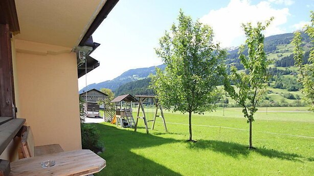 Un banc en bois se trouve à l’extérieur d’un bâtiment, offrant une vue sur un champ verdoyant, des arbres et une montagne en arrière-plan.