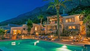 A poolside area with lounge chairs and a swimming pool, surrounded by a building with multiple balconies and a mountain backdrop. - Petite Fleur by Interhome (Jávea)