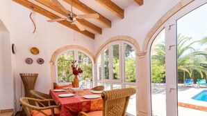 A dining area with a table set for four, surrounded by wicker chairs. There are arched windows with a view of a swimming pool and palm trees. - Villa Mariposa by Interhome (Jávea)