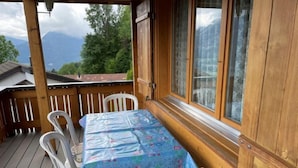 Une terrasse en bois avec une table recouverte d’une nappe à motifs, des chaises en plastique blanc et une vue sur les montagnes par une fenêtre.