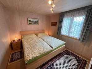 A wooden-paneled room with a bed, a nightstand, a window with curtains, and a painting on the wall.