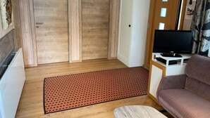 Un espace de vie compact doté d’un plancher en bois, d’un tapis rouge à motifs, d’un canapé, d’un téléviseur posé sur un meuble et d’une porte en bois.