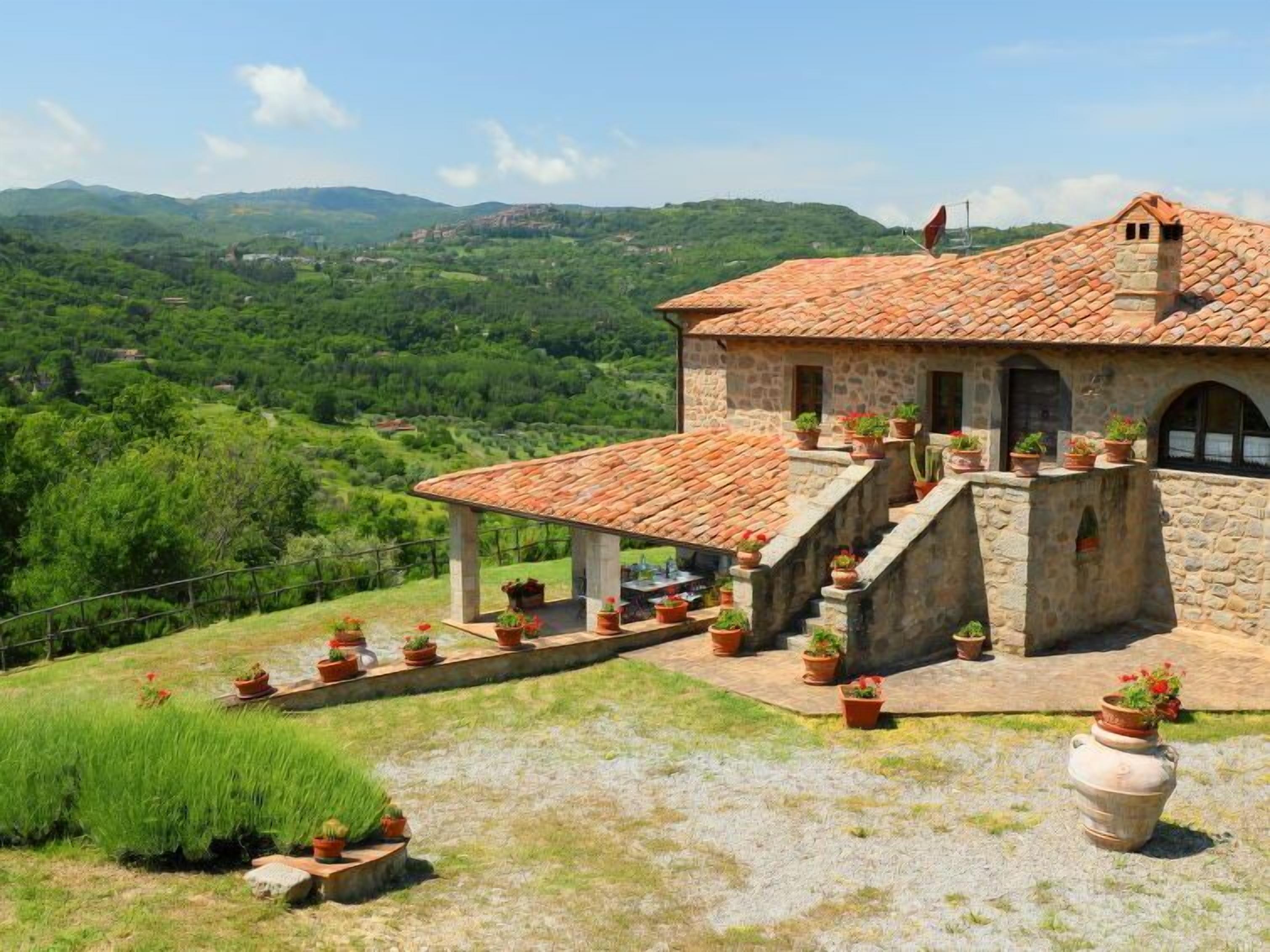 Un bâtiment en pierre avec un toit en tuiles, un patio couvert agrémenté de plantes en pot et une vue sur une vallée verdoyante.