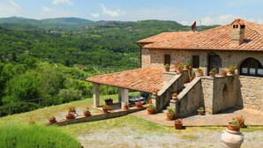Un bâtiment en pierre avec un toit en tuiles, un patio couvert agrémenté de plantes en pot et une vue sur une vallée verdoyante.