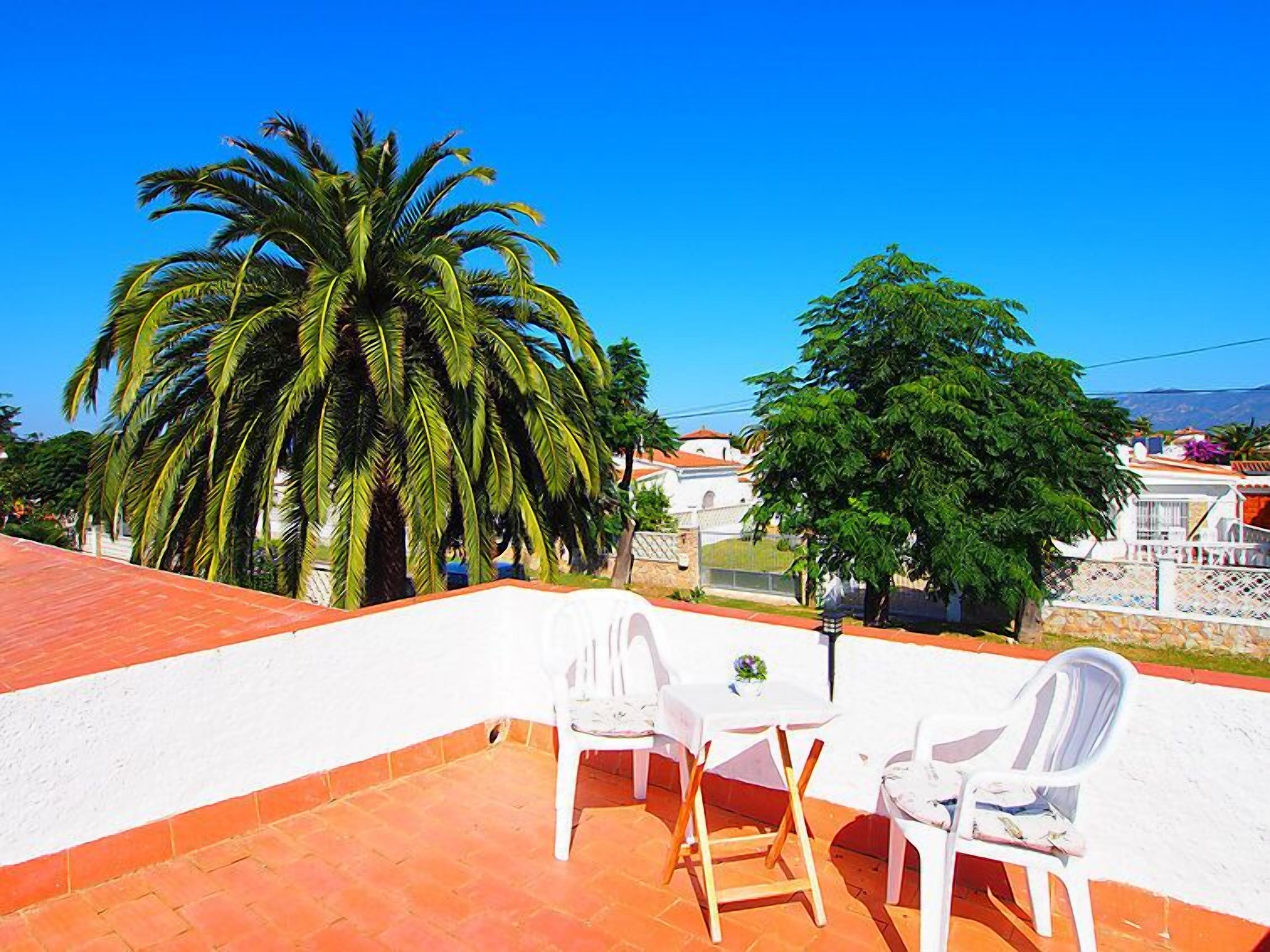 Une terrasse avec des chaises blanches et une table, entourée de palmiers et offrant une vue sur des maisons.