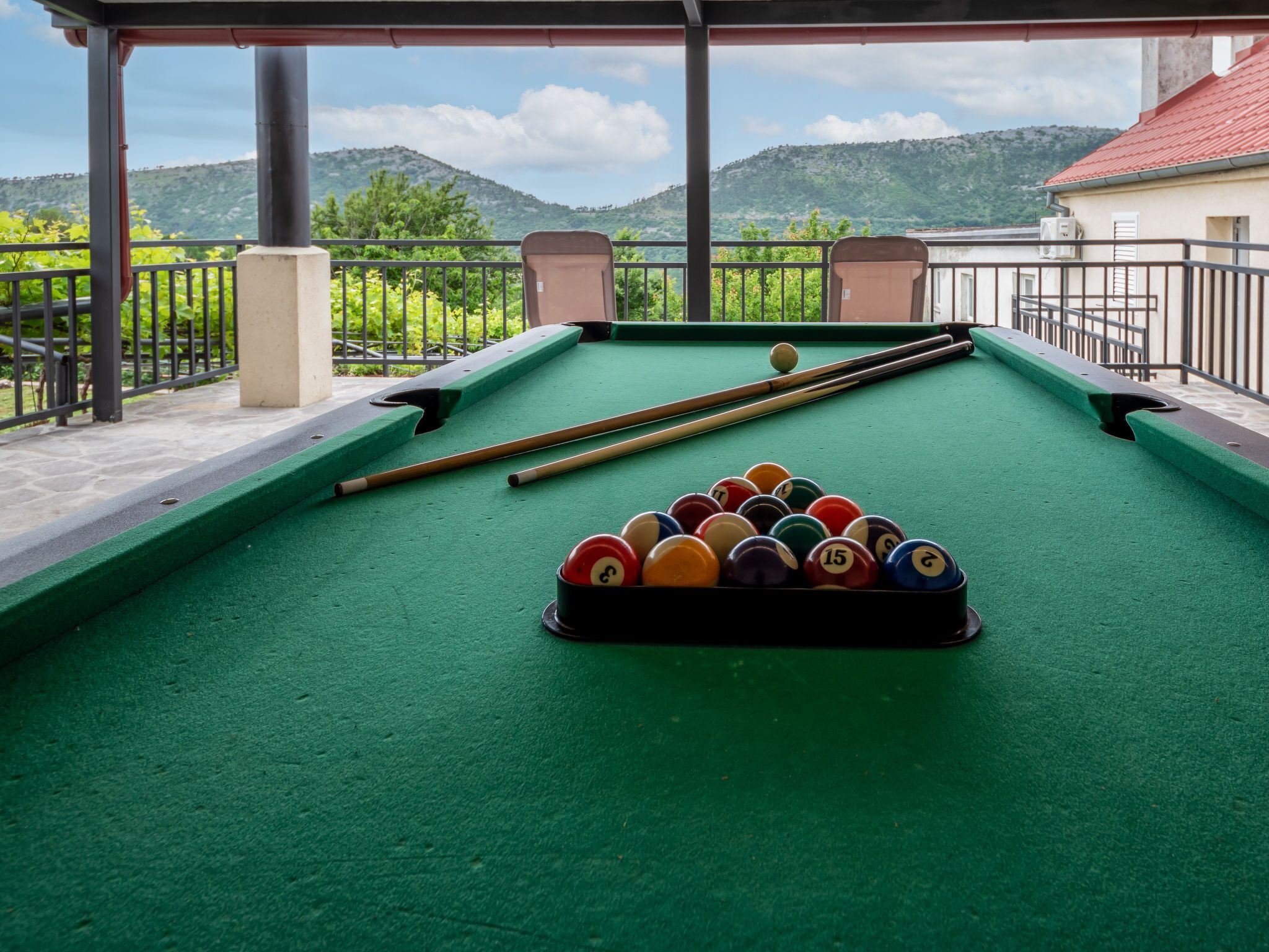 A pool table with balls and cues, set on a balcony with a view of green hills and a clear sky.