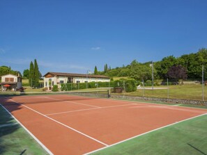 Ein Tennisplatz mit Netz, umzäunt und neben einem Gebäude mit Satteldach.