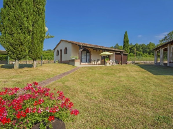 Una casa con porche cubierto, césped y jardín con flores rojas.
