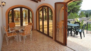 Un patio cubierto con una mesa y sillas, puertas de cristal arqueadas y vistas a un jardín y árboles.