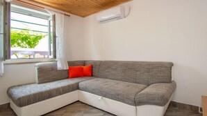 Un sofá de esquina con base blanca y cojines grises, techo de madera y ventana con vista a la vegetación.