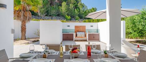 An outdoor dining area with a table set for six, a grill, and a view of a mountainous landscape.