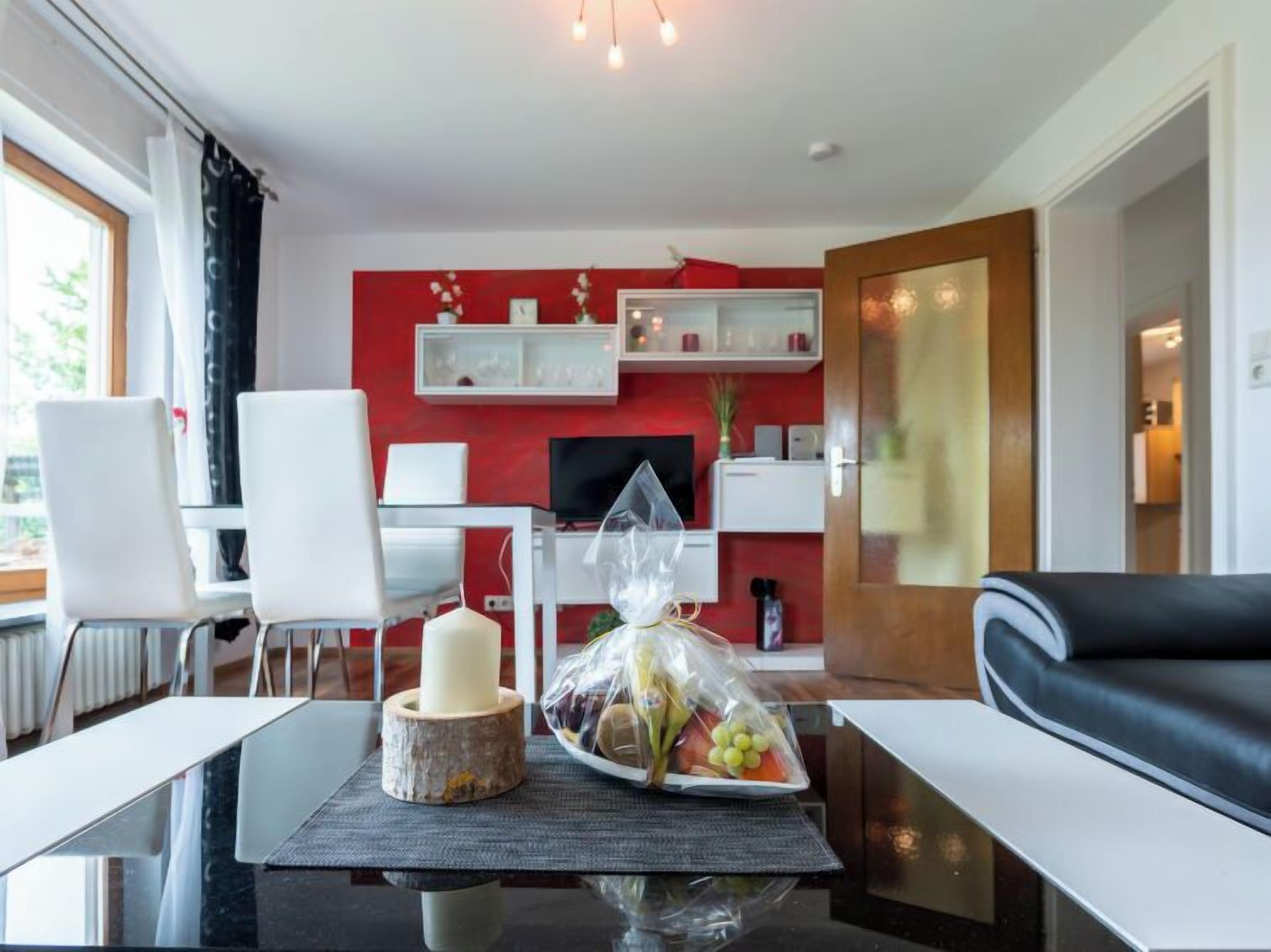 Ein modernes Wohnzimmer mit roter Akzentwand, einem schwarzen Ledersofa, einem Glastisch mit Kerze und Obst sowie einem weißen Schreibtisch mit Computer.