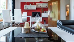 Una sala moderna con una pared de acento roja, un sofá de cuero negro, una mesa de centro de vidrio con una vela y fruta, y un escritorio blanco con una computadora.
