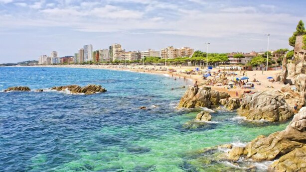 A coastal beach with clear blue waters, rocky formations, and a cityscape in the background.