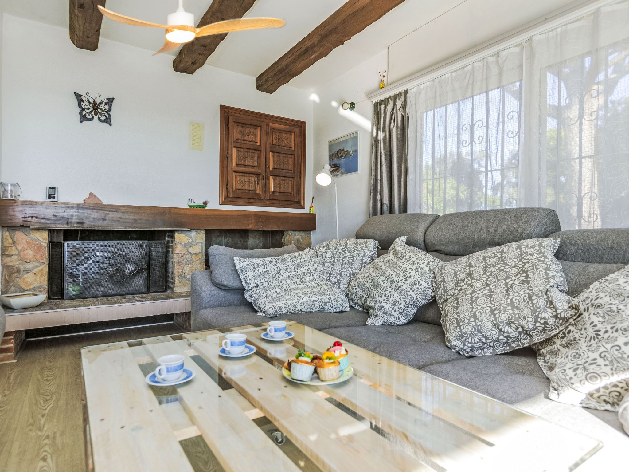 Ein Wohnzimmer mit Kamin, einer Couch mit gemusterten Kissen, einem Couchtisch mit Tassen und Kuchen sowie einem Deckenventilator.