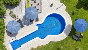 Vista aerea di una piscina dalla forma particolare a chitarra, circondata da un solarium in pietra con numerosi ombrelloni e lettini.