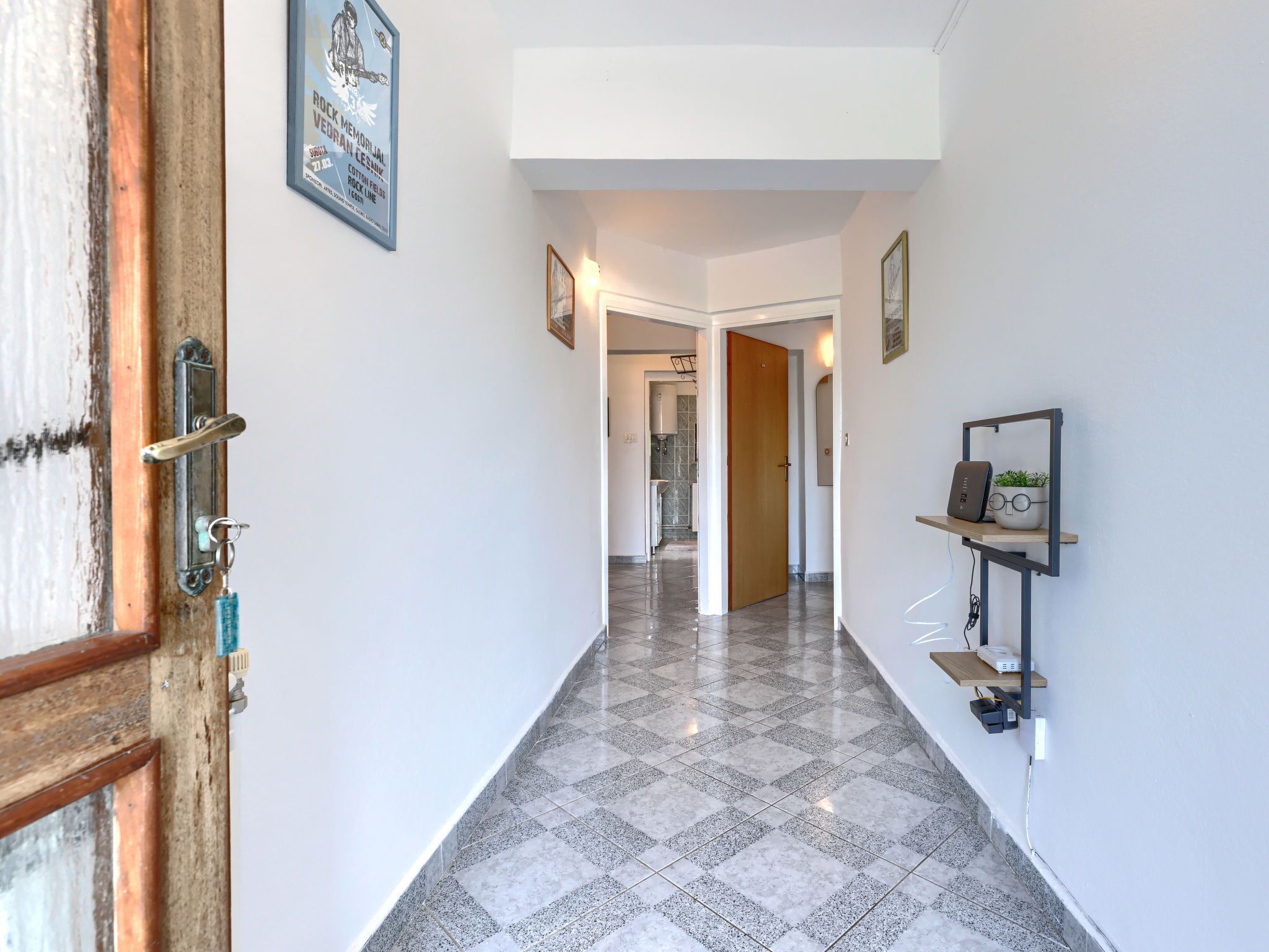 A hallway with a tiled floor, a wooden door, and a wall-mounted shelf with a plant and a speaker.