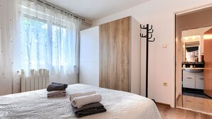 Una camera da letto con un letto grande, un termosifone, una finestra con tende leggere e un appendiabiti, con vista sul bagno.