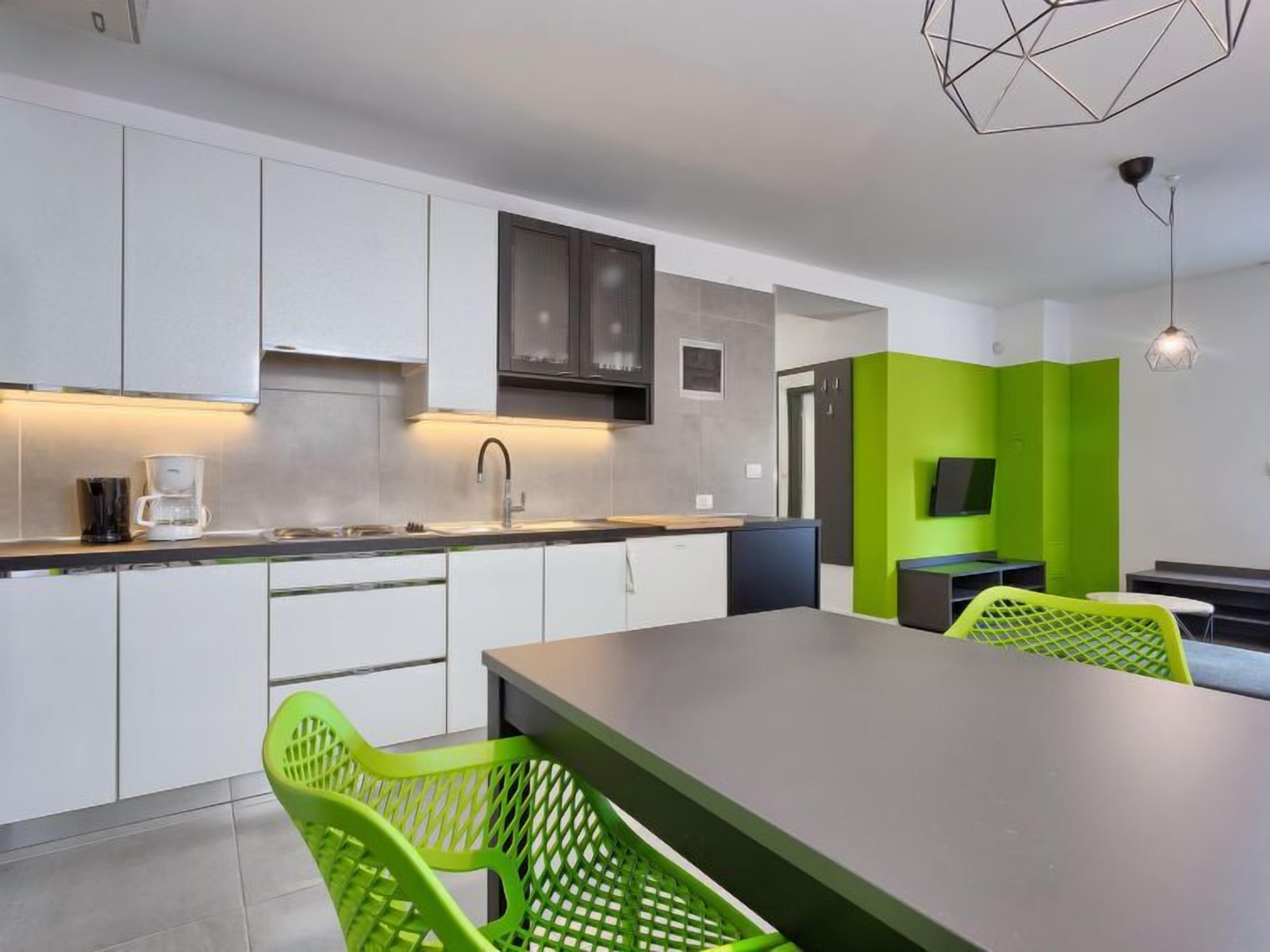 A modern kitchen with white cabinets, a grey countertop, and a large island with a grey top. There are two green chairs and a TV mounted on the wall.