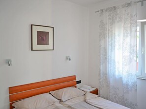 A bedroom with a wooden headboard, a framed picture on the wall, and a window with floral curtains. - Jadranka by Interhome (Rab/Rab)