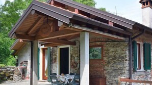 Un rústico edificio de piedra con un porche de madera, una mesa y sillas, y un muro de piedra con un cuadro.