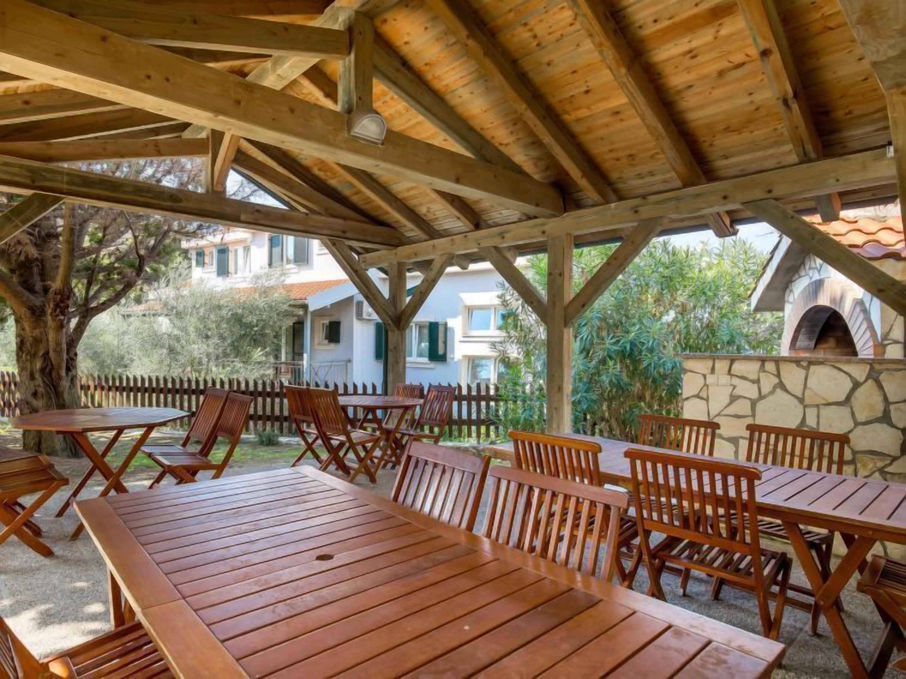A covered outdoor seating area with wooden tables and chairs, surrounded by a stone wall and greenery.