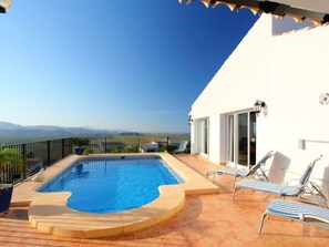 Une piscine avec vue sur les montagnes lointaines, un bâtiment blanc doté d’un balcon et de chaises longues.
