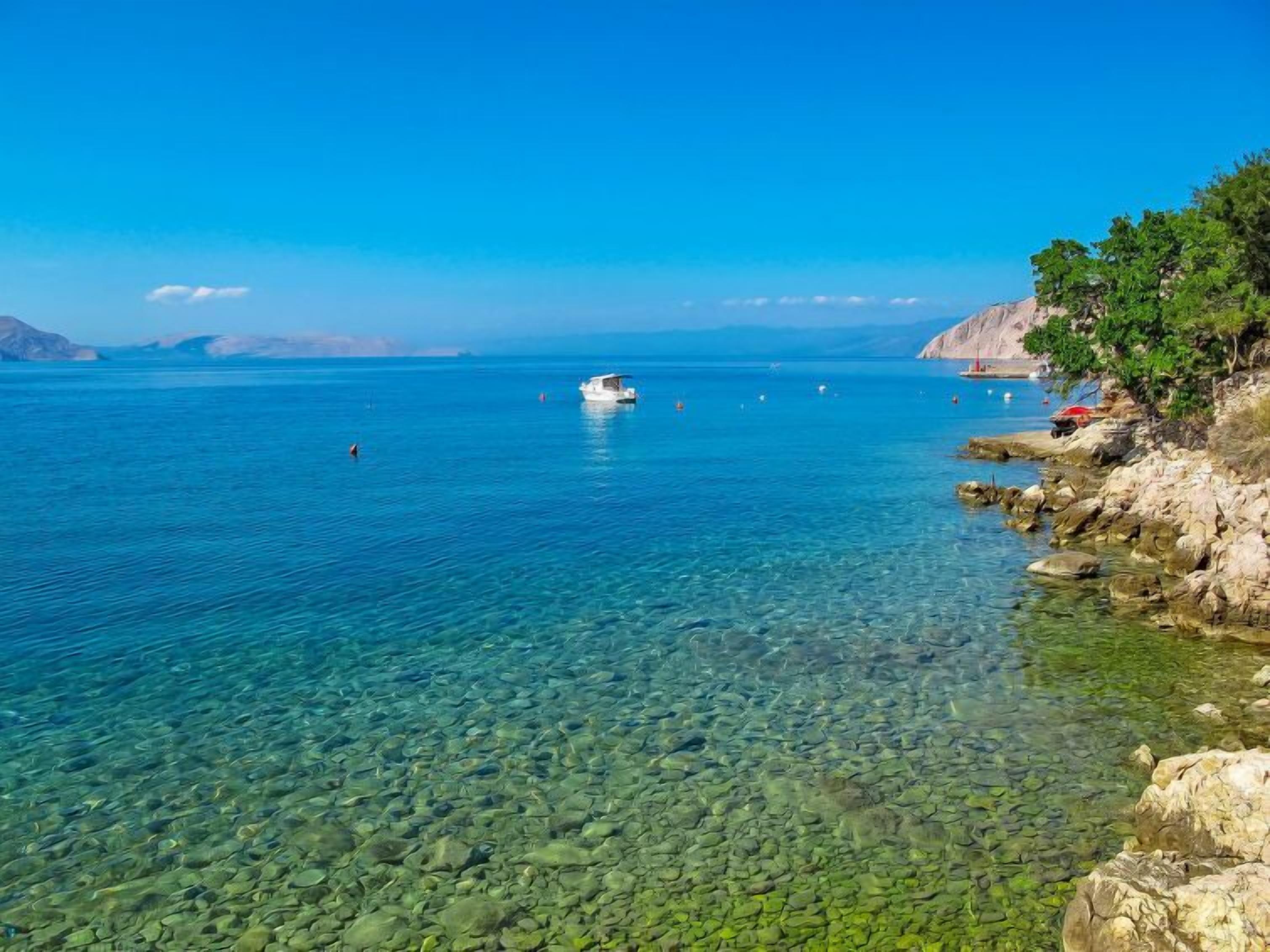 A clear coastal view with a boat on the water, rocky shoreline, and distant mountains.